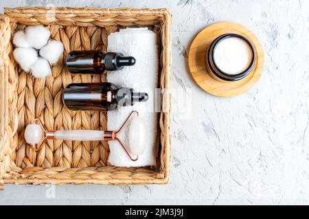 Draufsicht auf Flaschen aus dunklem Glas ohne Marken mit Hautpflegeprodukten in einem Öko-Korb. Gesicht Massage Roller und Baumwollblume. Gesichtsbehandlung Set für Home p Stockfoto