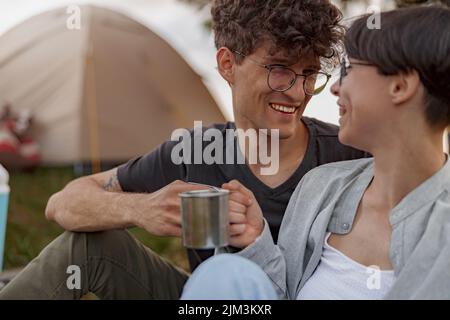 Porträt eines jungen fröhlichen Paares, das im Zelt im Freien sitzt, lacht und Kaffee in Tassen trinket. Stockfoto