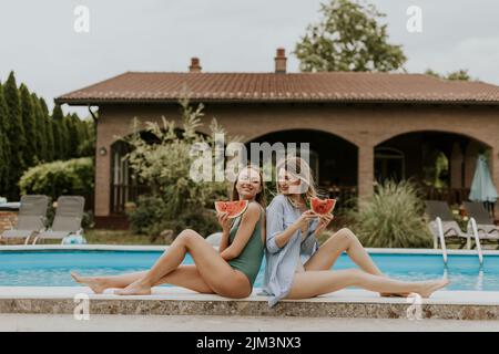Zwei niedliche junge Frauen sitzen am Pool und essen Wassermelone im Hinterhof des Hauses Stockfoto