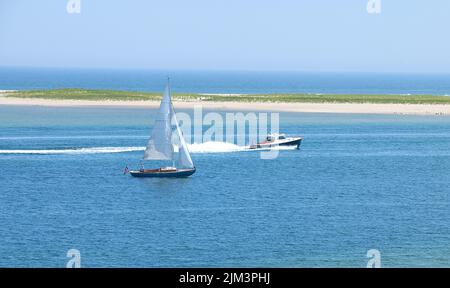 Ein Motorboot, das an einem Segelboot im Kanal vor Lighthouse Beach, Chatham, Massachusetts, auf Cape Cod, USA, vorbeifährt. Chatham Bar im Hintergrund Stockfoto