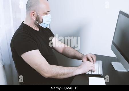Eine Nahaufnahme eines Mannes, der eine schwarze T-Short trägt und neben einem Computer und einer Maske auf seinem Gesicht sitzt Stockfoto