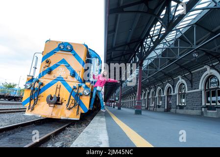 Ein Mädchen in einer rosa Jacke posiert für die Kamera aus einem Zug auf dem Bahnsteig des Dunedin Bahnhofs, Neuseeland Stockfoto
