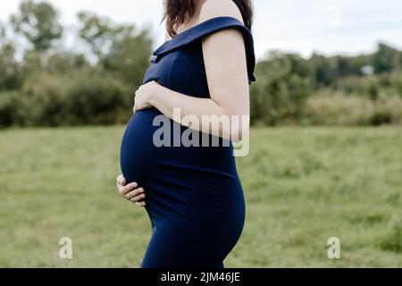 Eine junge, 38-wöchige Schwangere in einem blauen Kleid, das ihren Bauch hält Stockfoto