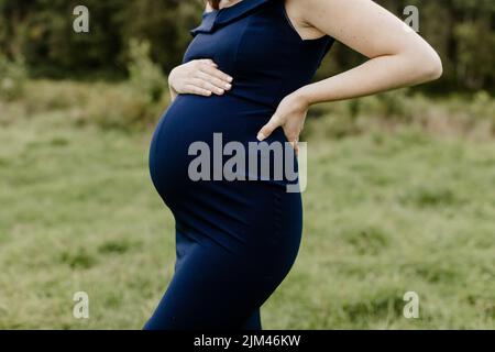 Eine junge, 38-wöchige Schwangere in einem blauen Kleid, das ihren Bauch hält Stockfoto