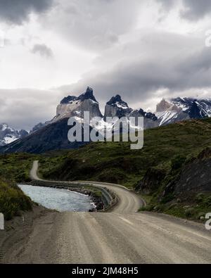 Eine vertikale Aufnahme einer Straße zwischen einem See und Grün gegen verschneite Berge an einem düsteren Tag Stockfoto