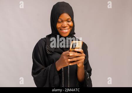 Eine afrikanisch-muslimische lächelnde, aufgeregt Dame, die ihr Telefon benutzte und auf grauem Hintergrund isoliert war Stockfoto