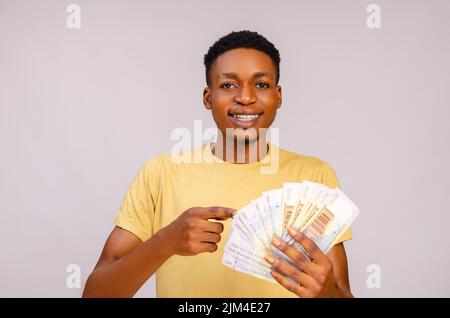 Eine Nahaufnahme eines jungen afrikanischen Mannes, der Papierrechnungen in der Hand hält und lächelnd auf sie zeigt Stockfoto