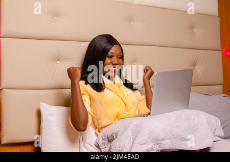 Eine Nahaufnahme einer schönen schwarzen Frau, die in einem Bademantel im Bett lag und aufgeregt auf ihren Laptop schaute Stockfoto