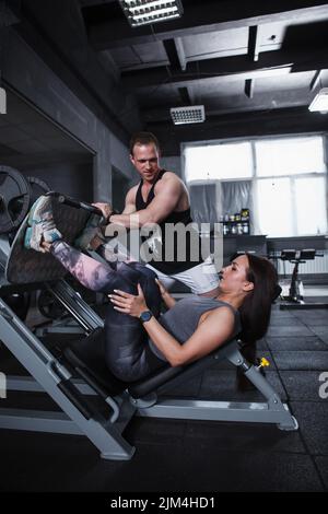 Vertikale Aufnahme eines professionellen Personal Trainers, der seine Kundin beim Training mit einem Fitnessgerät für Beinpresse unterstützt Stockfoto