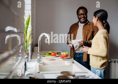 Schwarzer bärtiger Mann lächelt, während er mit seiner Tochter zu Hause Sandwiches zubereitet Stockfoto