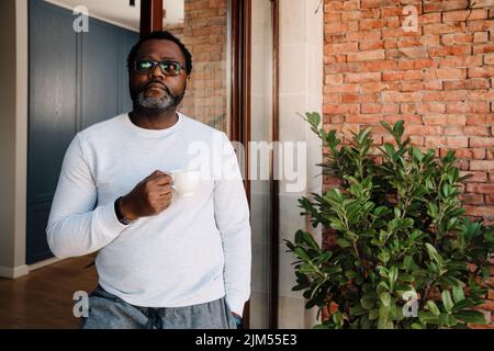 Schwarzer bärtiger Mann, der Kaffee trinkt, während er zu Hause in der Tür steht Stockfoto