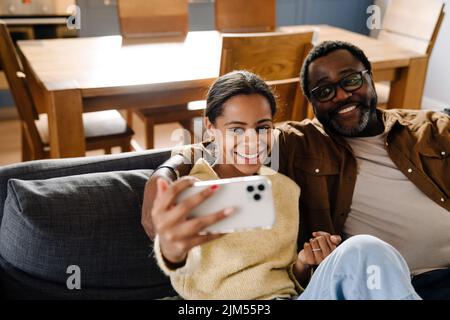 Das schwarze Mädchen und ihr Vater machen Selfie auf dem Handy, während sie zu Hause auf dem Sofa sitzen Stockfoto