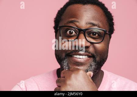 Schwarzer bärtiger Mann in einer Brille lächelt und blickt isoliert auf einen rosa Hintergrund auf die Kamera Stockfoto