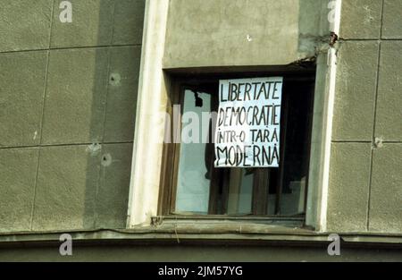 Bukarest, Rumänien, Januar 1990. Ein Plakat, das auf dem Universitätsplatz hängt, sagt 'Freiheit und Demokratie in einem modernen Land', Tage nach der antikommunistischen Revolution vom Dezember 1989. An der Wand des Gebäudes sind Einschusslöcher sichtbar. Stockfoto