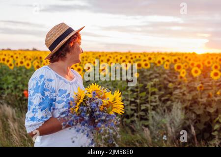 Ältere Frau hält bei Sonnenuntergang im Sommerfeld einen Strauß gelber Sonnenblumen. Frau mittleren Alters bewundert die Landschaft Stockfoto