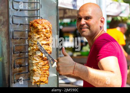 Koch schneidet mit Dönermesser traditionelles türkisches Dönerfleisch und zeigt Daumen nach oben. Shawarma oder Gyros. Türkischer, griechischer oder mittelöstlicher arabischer Stil Stockfoto