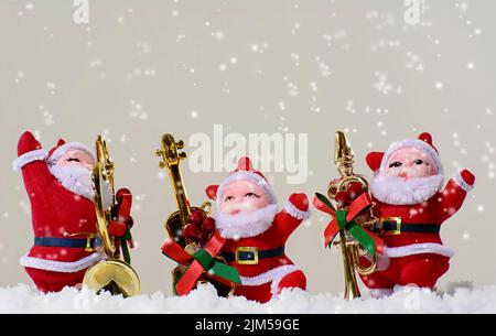 Die Schneeflocken fallen über drei kleine Figuren des Weihnachtsmannes mit Musikinstrumenten auf weißem Hintergrund Stockfoto