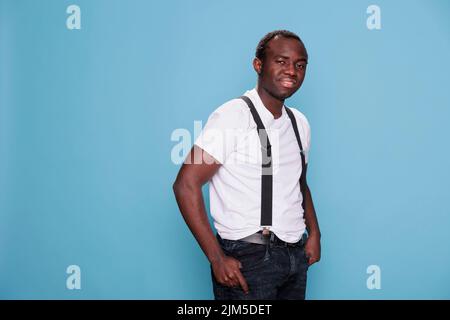 Selbstbewusster Mann, der vor der Kamera posiert, während er auf blauem Hintergrund steht. Lächelnd freundlich aussehende junge Erwachsene Person mit Mode-Stil tragen weißes T-Shirt und Hosenträger. Studioaufnahme Stockfoto