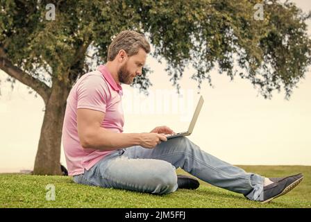 Konzentrierter Geschäftsmann mit Laptop zum Bloggen auf grünem Gras, arbeiten online Stockfoto