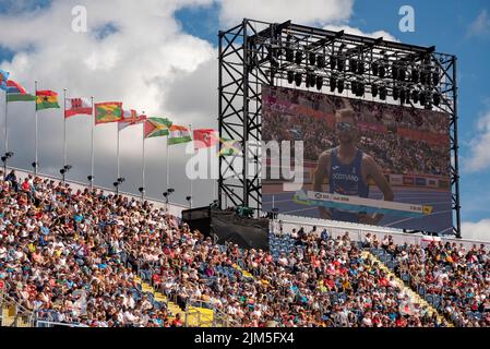 Birmingham, Großbritannien, 4.. August 2022. Zuschauer im Alexander Stadium, Birmingham, genießen die Leichtathletik bei den Commonwealth Games 2022. Flaggen der Commonwealth-Staaten fliegen über das Stadion. Josh Kerr aus Schottland, auf der Leinwand gesehen, qualifizierte sich für die Männer 1500m. Quelle: Martin Anderson/Alamy Live News Stockfoto