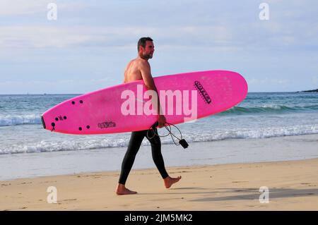 Mann läuft mit einem rosafarbenen Surfbrett entlang des Gwihian Beach, Cornwall, England. Stockfoto