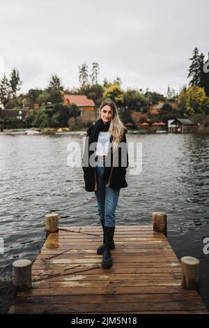 Junge blonde, warme Frau, die an einem bewölkten Nachmittag auf einem hölzernen Dock in einem schönen See steht. Speicherplatz kopieren Stockfoto