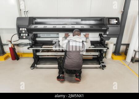 Der Bediener des Technikers wechselt die Papierrolle auf einem großen hochwertigen Industriedrucker und Plottergerät im digitalen Druckereibüro Stockfoto