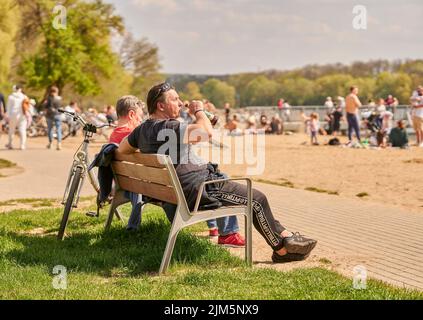 Ein Mann, der auf einer Holzbank sitzt und an einem sonnigen Frühlingstag in Posen, Polen, Bier trinkt Stockfoto