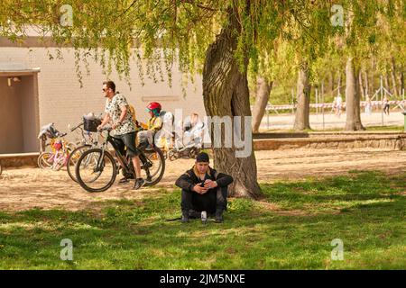 Ein Mann mit Telefon sitzt an einem sonnigen Frühlingstag unter dem Baum in Posen, Polen Stockfoto