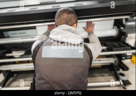 Der Bediener des Technikers wechselt die Papierrolle auf einem großen hochwertigen Industriedrucker und Plottergerät im digitalen Druckereibüro Stockfoto