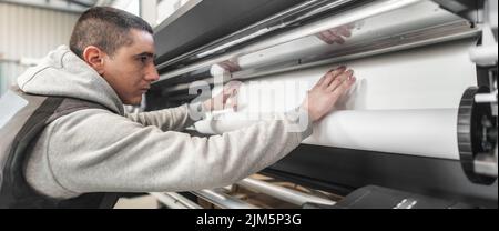 Der Bediener des Technikers wechselt die Papierrolle auf einem großen hochwertigen Industriedrucker und Plottergerät im digitalen Druckereibüro Stockfoto