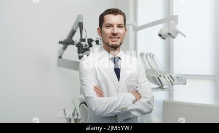 Glücklicher, männlicher Zahnarzt in weißem Labormantel, der mit gekreuzten Händen posiert und die Kamera anschaut Stockfoto