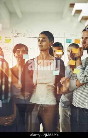 Eine Gruppe von Marketing-Profis oder Designern, die Ideen zum Brainstorming anstellen und während des Meetings auf ein transparentes Brett mit Haftnotizen schreiben. Vielfältiges Team von Stockfoto