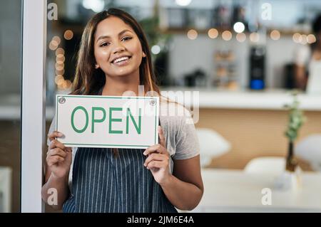 Es war immer mein Traum, dieses kleine Café zu eröffnen. Porträt einer jungen Frau, die ein offenes Schild in einem Café hält. Stockfoto