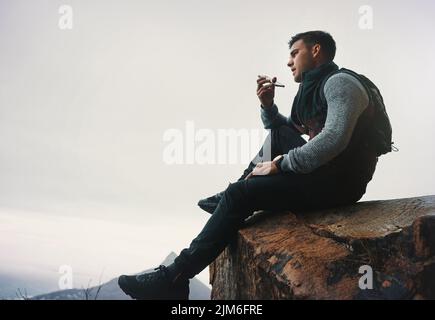 Ein junger Mann, der auf einem Handy spricht, während er auf einer Bergklippe sitzt. Stockfoto