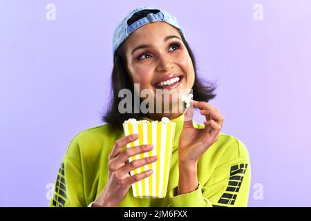 Die einprägsamsten Filme wirken nachhaltig. Studioaufnahme einer schönen jungen Frau, die vor purpurem Hintergrund Popcorn isst. Stockfoto