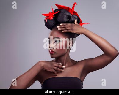 Make-up ist meine Kunst - mein Gesicht ist die Leinwand. Studioaufnahme einer schönen jungen Frau, die asiatisch inspiriertes Make-up trägt und mit Origami gegen ein Grau posiert Stockfoto