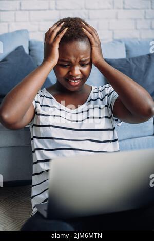 Afro-Frau, die den Kopf in den Händen hält, aufgeregt, besorgt über Online-Probleme. Schwarzes Mädchen, das aus der Ferne arbeitet, Freiberufler. Stockfoto
