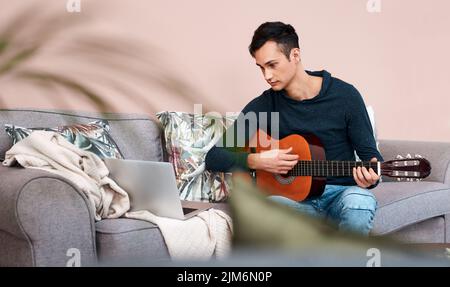Lockdown richtig gemacht. Ein junger Mann, der einen Laptop benutzt, während er zu Hause Gitarre spielt. Stockfoto