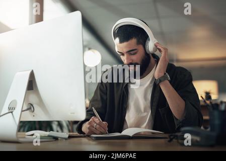 Gute Musik hält die kreativen Säfte fließen. Ein junger Geschäftsmann in einem Notebook schreiben und mit einem Computer mit Kopfhörern während eines späten Nachts Stockfoto