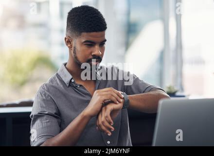 Ein junger Geschäftsmann überprüft die Zeit, während er an einem Laptop in einem Büro arbeitet. Stockfoto