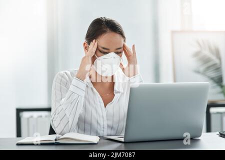 Eine maskierte junge Geschäftsfrau, die gestresst aussieht, während sie an ihrem Schreibtisch in einem modernen Büro arbeitet. Stockfoto