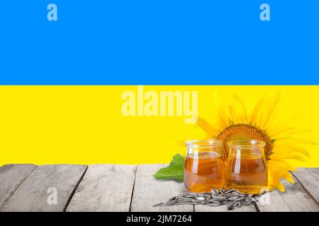 Gläser Sonnenblumenöl und Samen auf Holztisch gegen die Flagge der Ukraine Stockfoto