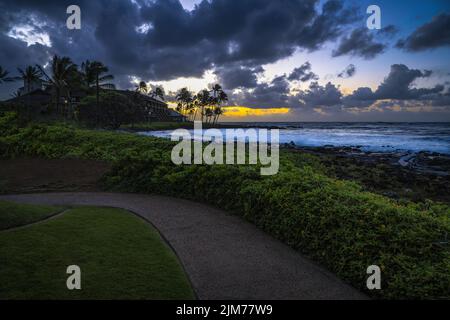 AM FRÜHEN ABEND BRICHT EIN WARMER SONNENUNTERGANG DURCH DUNKLE WOLKEN IN DER NÄHE DES PILI MAI RESORTS AUF KAUAI HAWAII Stockfoto