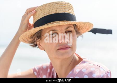 Nachdenkliche verheiratete Frau mit grauen Haaren in einem rosa Kleid mit weißen Tupfen, die den Bootsfahrer mit der Hand halten, die der Wind von ihrem Kopf zu blasen versucht. Stockfoto