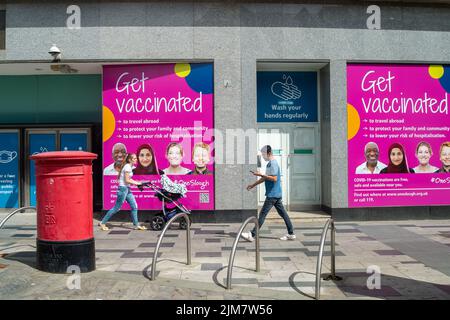 Slough, berkshire, Großbritannien. 4.. August 2022. In der Slough High Street laufen die Käufer an den neuen neonrosa Hinweisschildern vorbei, an die man sich geimpft lassen kann. Der Gemischte Ausschuss für Impfungen und Impfungen (JCVI) gab kürzlich bekannt, dass alle über 50 Jahre zu den im Herbst im Rahmen der Pläne, den Schutz vor Atemwegsviren vor dem Winter zu erhöhen, angebotenen COVID-19-Impfstoffen und Grippejabs gehören werden. Quelle: Maureen McLean/Alamy Live News Stockfoto