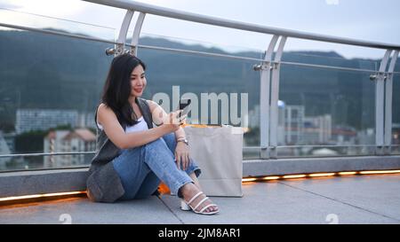 Hübsche junge Frau mit Einkaufstaschen, die bei Sonnenuntergang vor dem Hintergrund der Stadtlandschaft auf der Terrasse sitzt Stockfoto