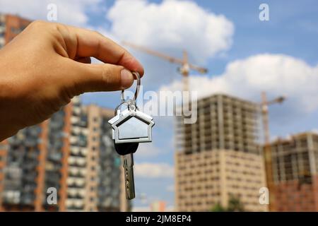 Immobilienmakler, Schlüsselanhänger in Form eines Hauses und Schlüssel in männlicher Hand auf dem Hintergrund von Baukräne und Neubauten. Byung Wohnung oder Miete Stockfoto