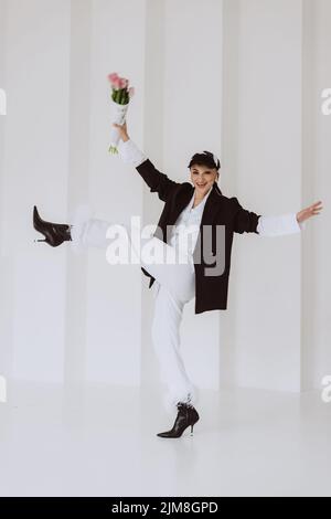 Eine fröhliche, stilvolle Frau in einem modischen Outfit, ein hellweißer Anzug mit Federn, eine schwarze Jacke, eine Baseballmütze und mit einem Blumenstrauß in ihrem h Stockfoto