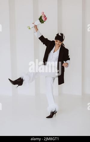 Eine fröhliche, stilvolle Frau in einem modischen Outfit, ein hellweißer Anzug mit Federn, eine schwarze Jacke, eine Baseballmütze und mit einem Blumenstrauß in ihrem h Stockfoto